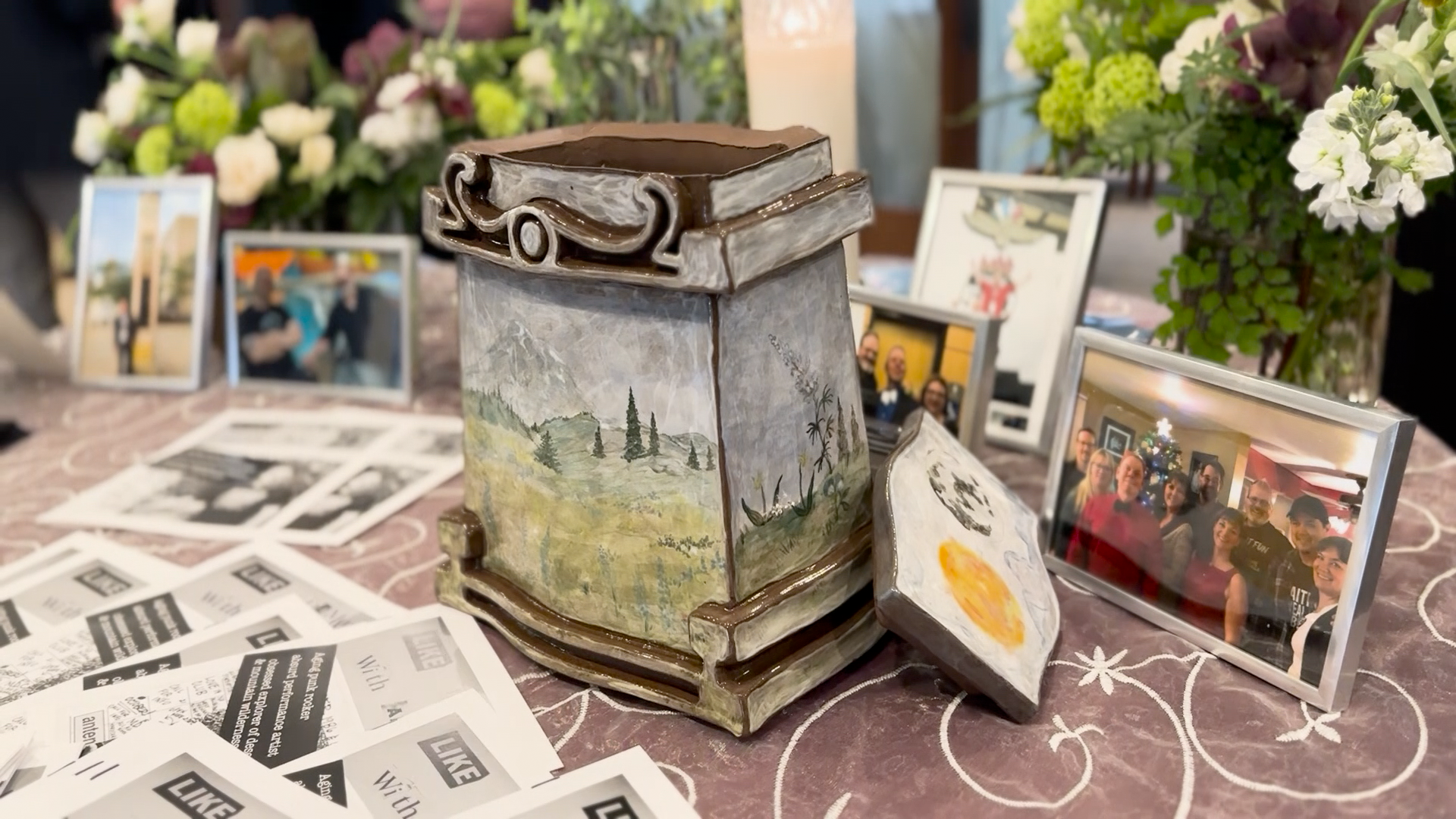 A decorative urn with a landscape design sits on a table surrounded by framed photographs and floral arrangements, capturing memories of a loved one.