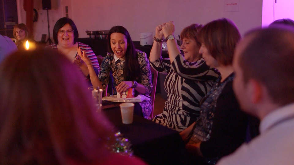 Group of adults celebrating around a table indoors, some raising hands and smiling. A lit candle and drinks on the table.