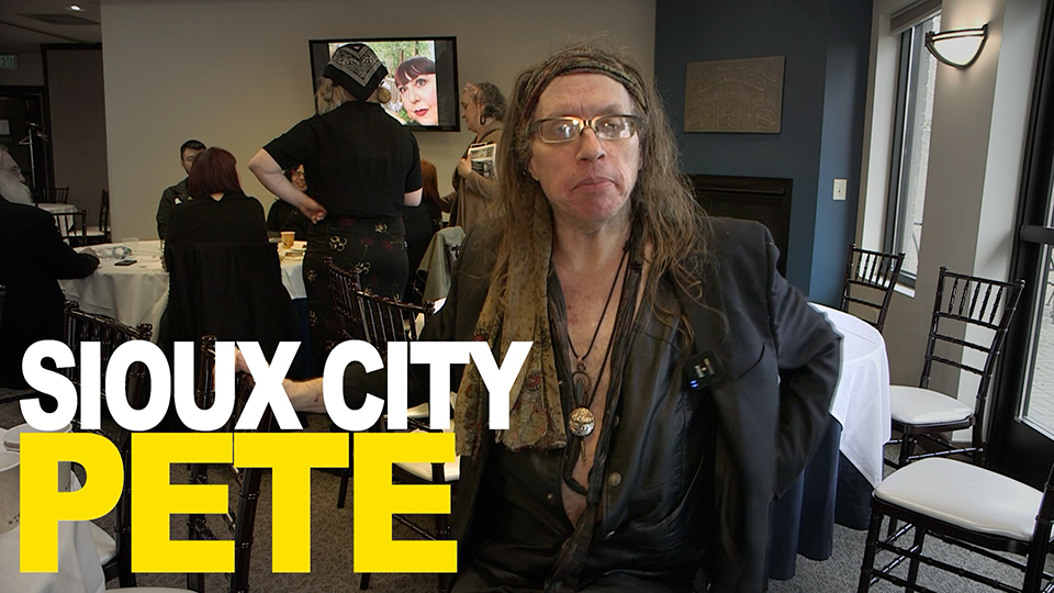 Person with long hair and glasses in a dark blazer seated at a dining table in a banquet room; large white and yellow text 'SIOUX CITY PETE' overlays the left side of the image.
